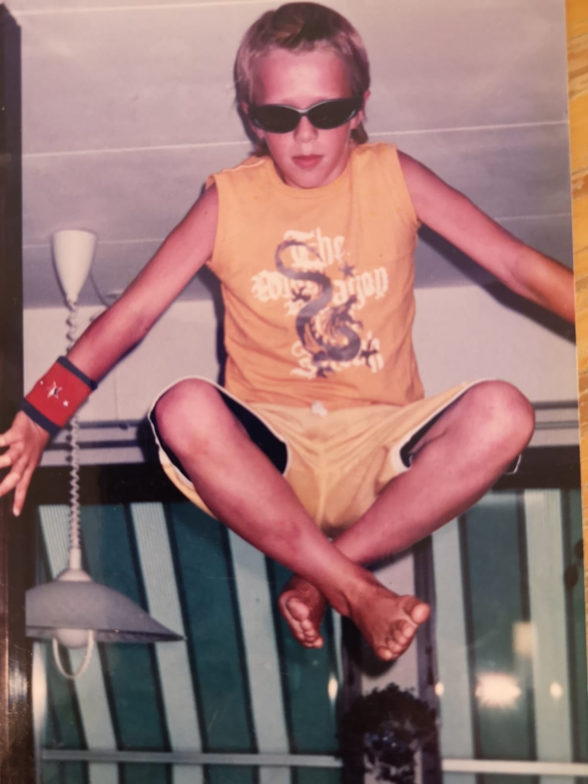 Birk as a kid, holding himself up in a doorway with his hands, legs crossed, in sunglasses and a dragon shirt